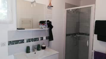Een badkamer met een wit wastafel, spiegel en douche. Er staan groene flessen op het aanrecht en een zwarte handdoek hangt aan de muur.