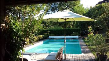 Relax by the serene pool surrounded by lush greenery and shaded by a large umbrella.