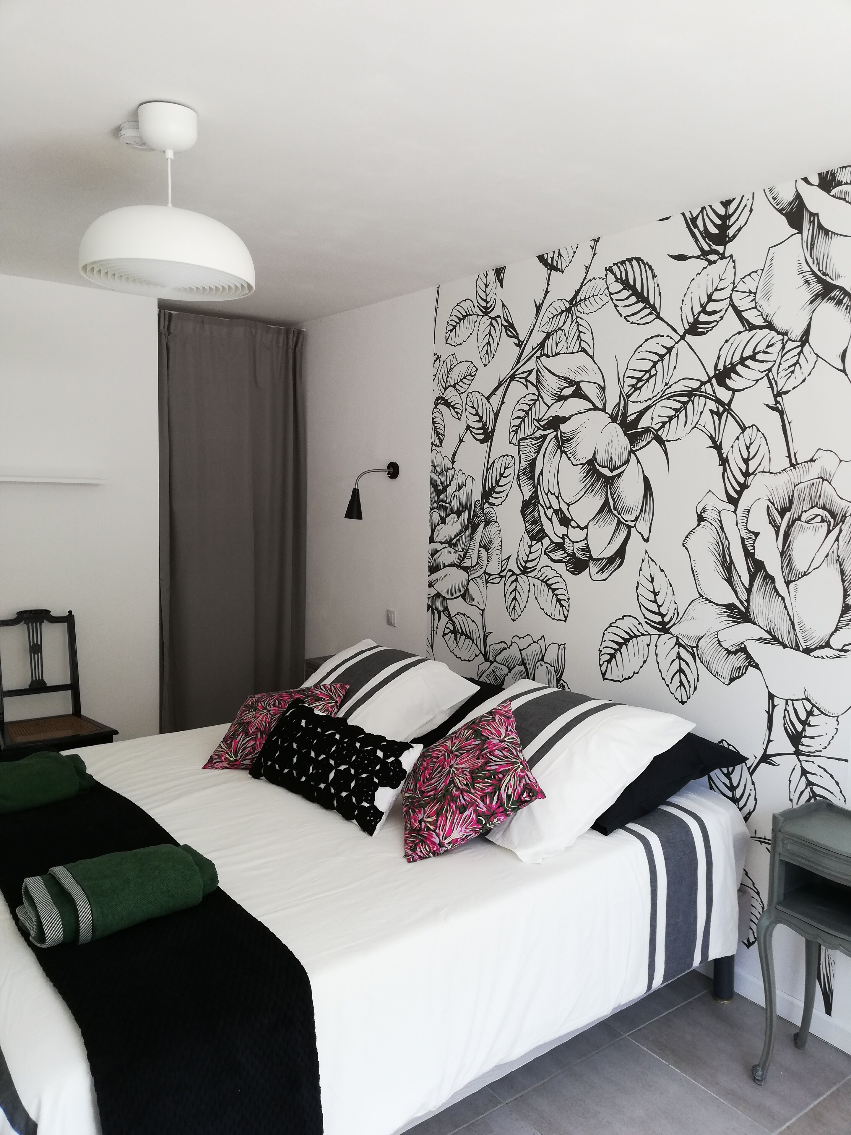 A bedroom with a large bed, a bedside table, a chair, a hanging light fixture, and a floral wallpaper.
