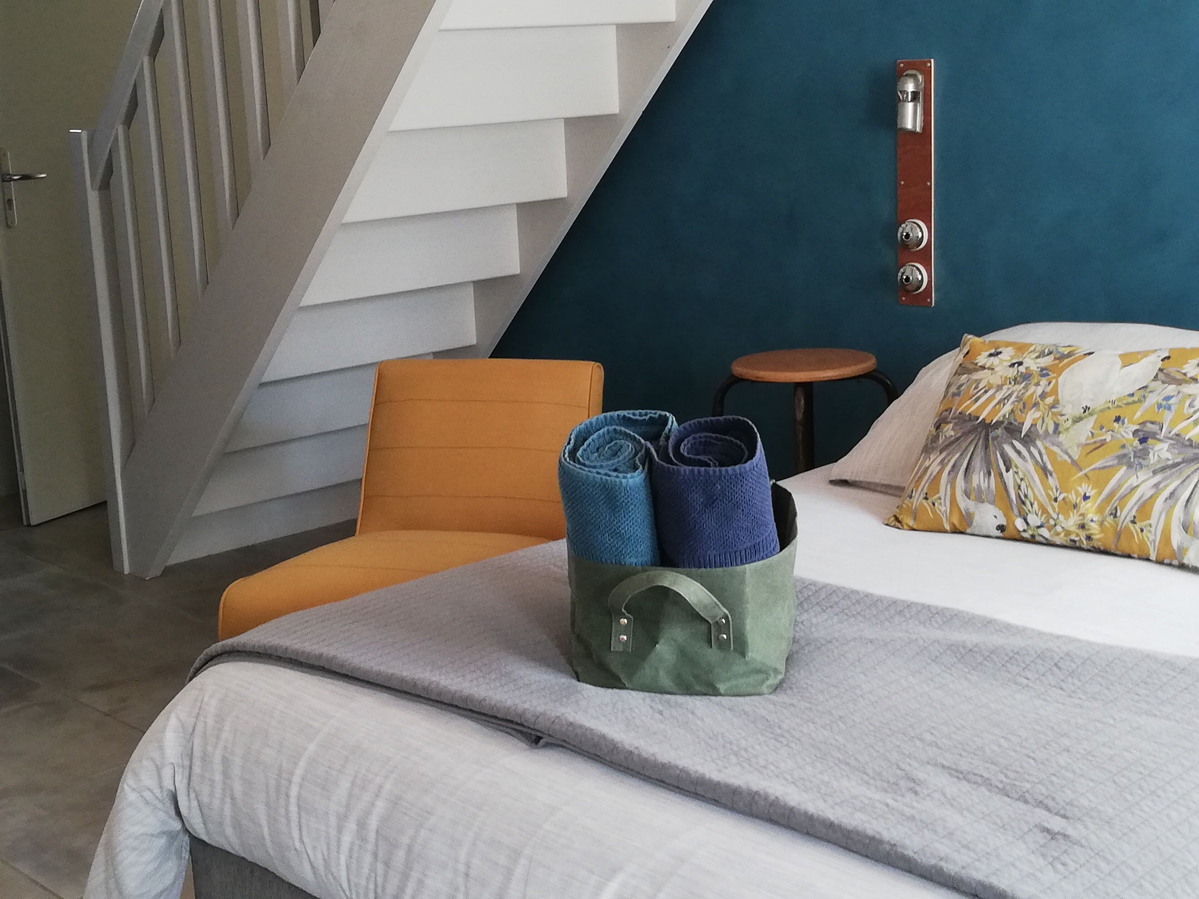 A hotel room with a bed, a green bag with rolled towels, an orange chair, a small stool, and a staircase.