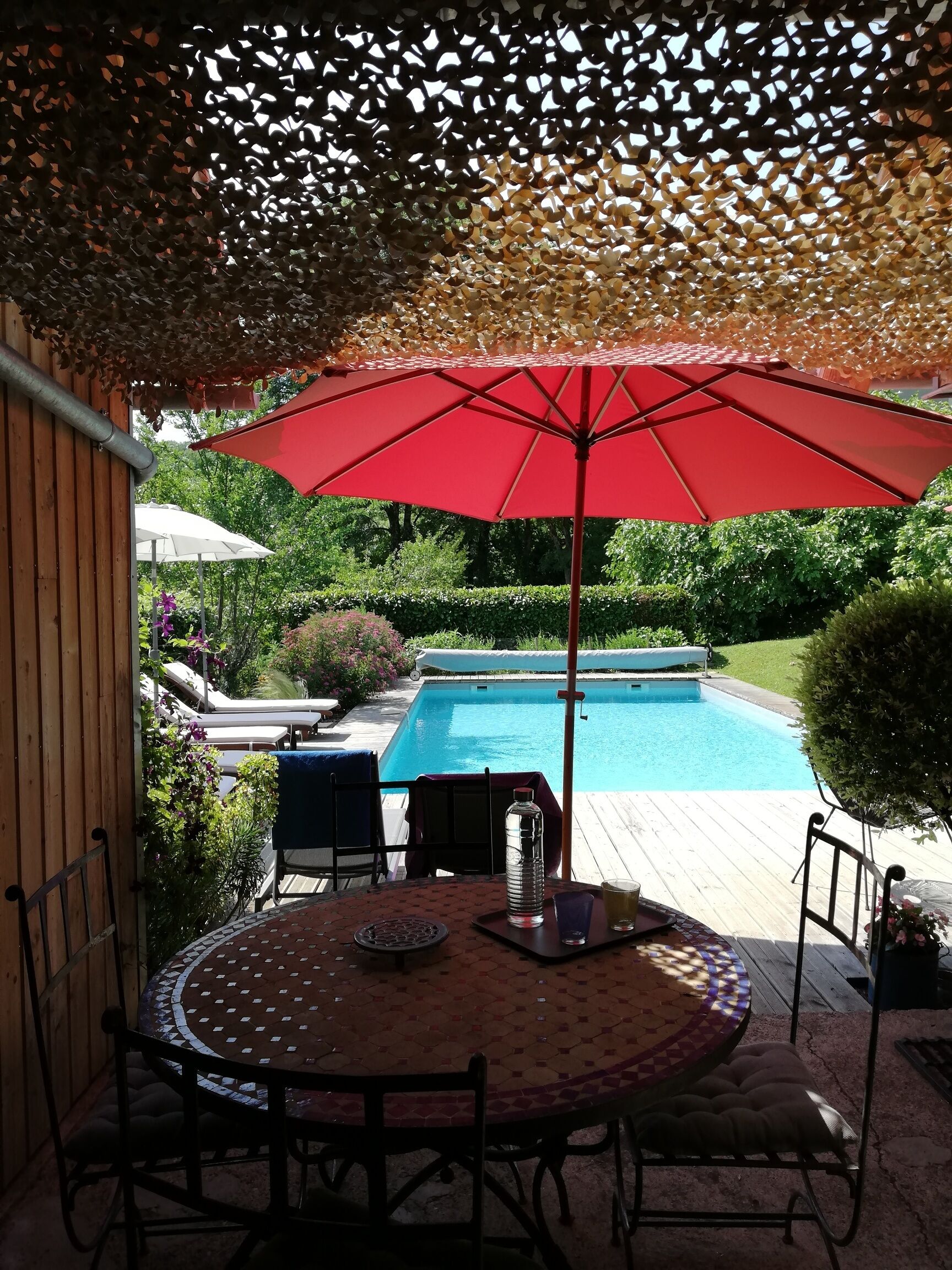 A poolside patio with a round table, chairs, a red umbrella, and a pool.