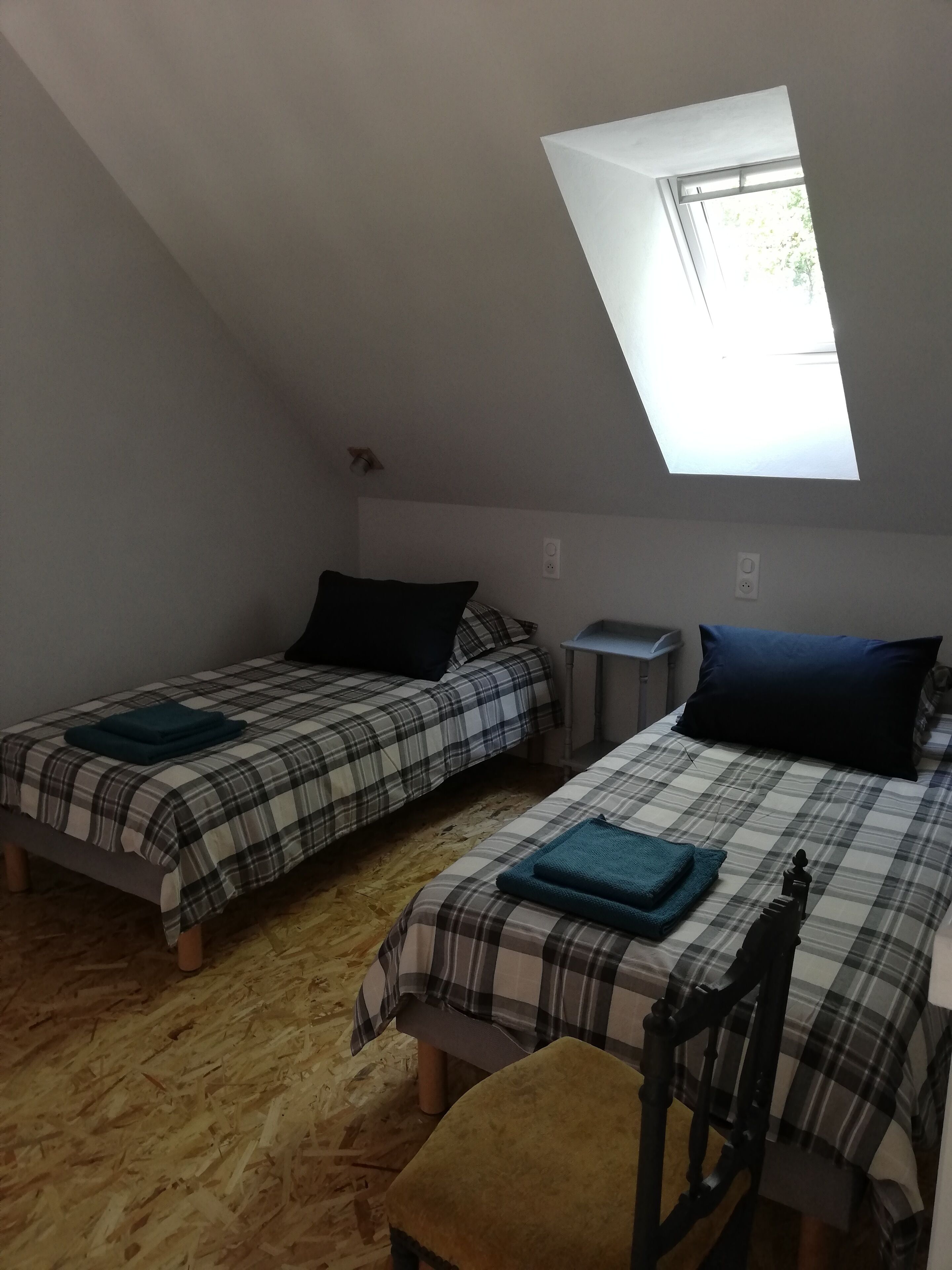 A dormitory room with two beds, each with a plaid bedspread and a dark pillow. There is a small table and a chair. The room has a sloped ceiling with a skylight.