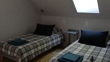A dormitory room with two beds, each with a plaid bedspread and a dark pillow. There is a small table and a chair. The room has a sloped ceiling with a skylight.