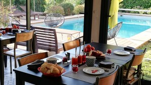Un espace repas avec vue sur une piscine, des sièges d’extérieur et une table dressée avec de la nourriture et des boissons.