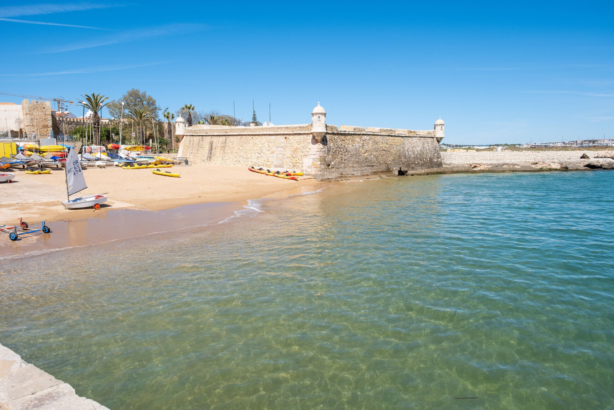 Una zona costiera con spiaggia sabbiosa, acque limpide e un muro di pietra con un faro.