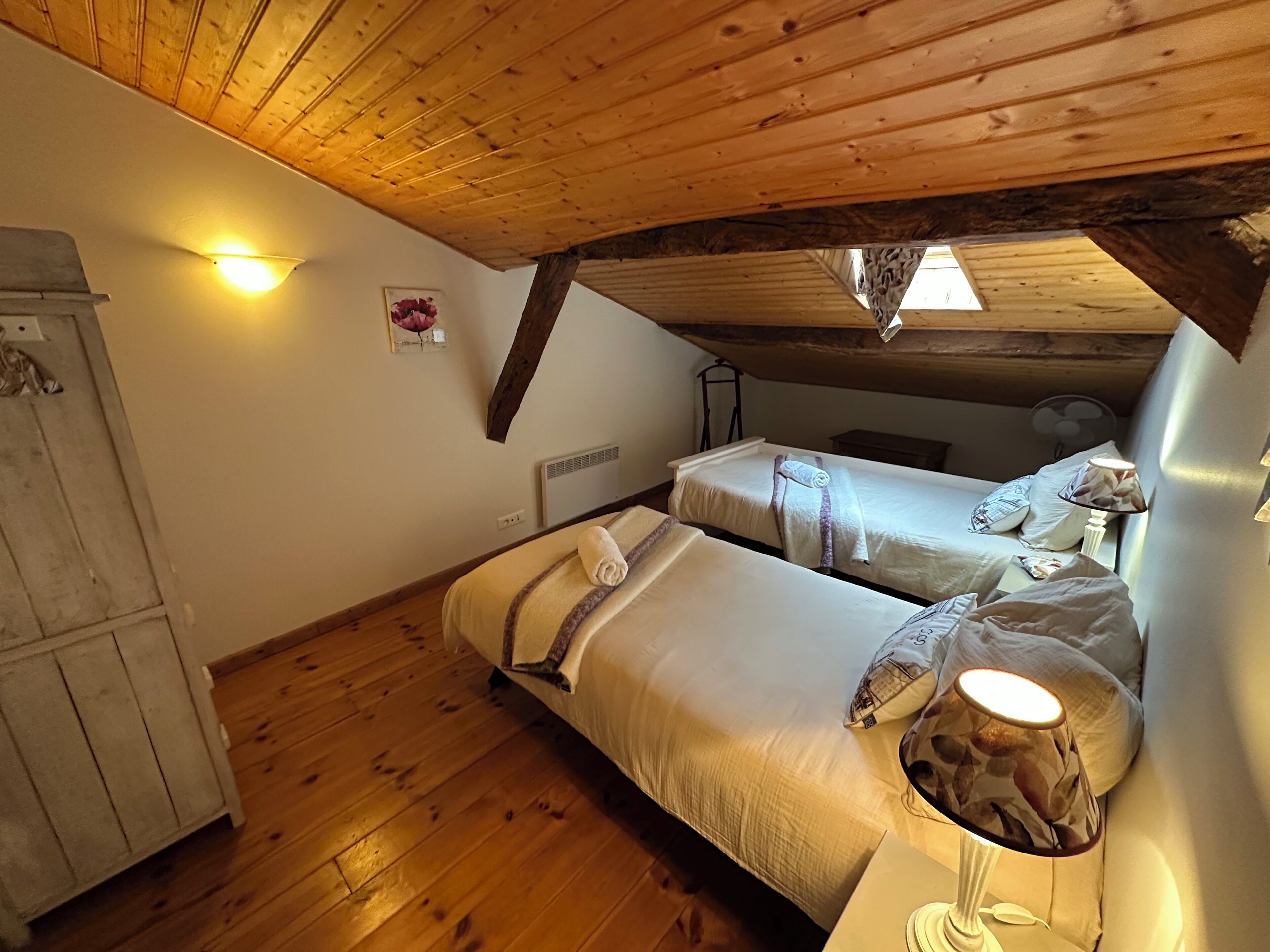 Un dormitorio acogedor en el ático con vigas de madera, dos camas, un armario y una mesita con lámpara.