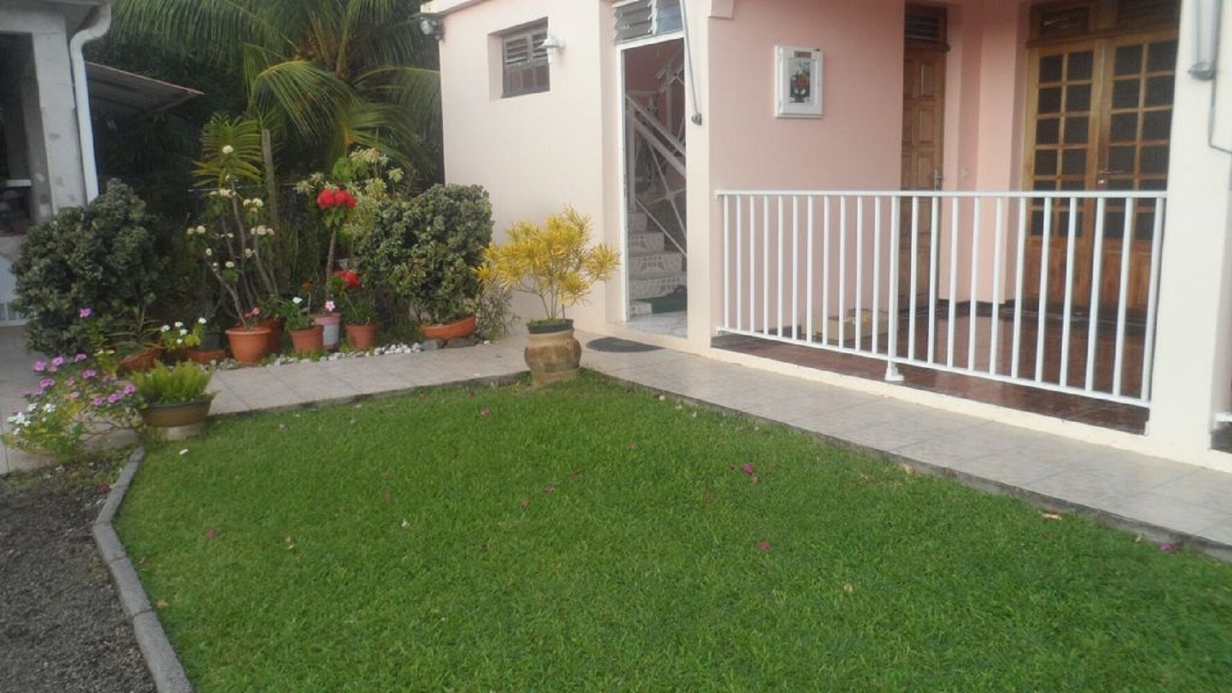 Un césped bien cuidado con plantas en macetas y una cerca blanca frente a un edificio con una pared rosa y una ventana.