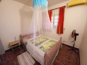 Une chambre à coucher comprenant un lit, une table de chevet, un ventilateur sur pied, une fenêtre avec des rideaux rouges et un appareil de climatisation.