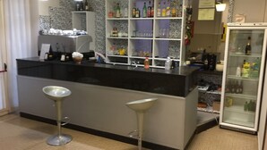 A bar area with a black countertop, two bar stools, shelves with bottles, and a refrigerator. - Hotel Cuba Rimini (Rimini)