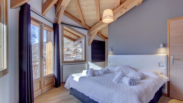 Een slaapkamer met een groot bed, houten balken, een raam met gordijnen en uitzicht op een bergachtig gebied.