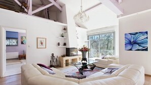 A spacious living room with a large white sofa, a glass coffee table, and a chandelier. There is a television on a wooden stand, and a window with a view of trees.