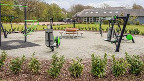 Uma área de ginástica ao ar livre com equipamentos de exercício, um banco de madeira e um edifício ao fundo.