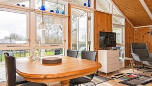 Un comedor amplio con una mesa y sillas de madera, un televisor en un mueble, y ventanas grandes que ofrecen vistas al exterior.