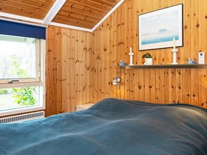 A room with wooden walls, a bed with a blue blanket, a window with a view of greenery, a wall-mounted shelf with decorative items, and a framed picture above the shelf.