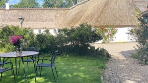 Un edificio con techo de paja y fachada blanca, rodeado de un jardín cuidado con una mesa redonda y sillas, y un camino de grava que lleva a la entrada.