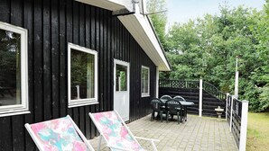 A black wooden building with a covered patio area featuring two lounge chairs and a table. - 4 Person Holiday Home in Ansager (Ansager)