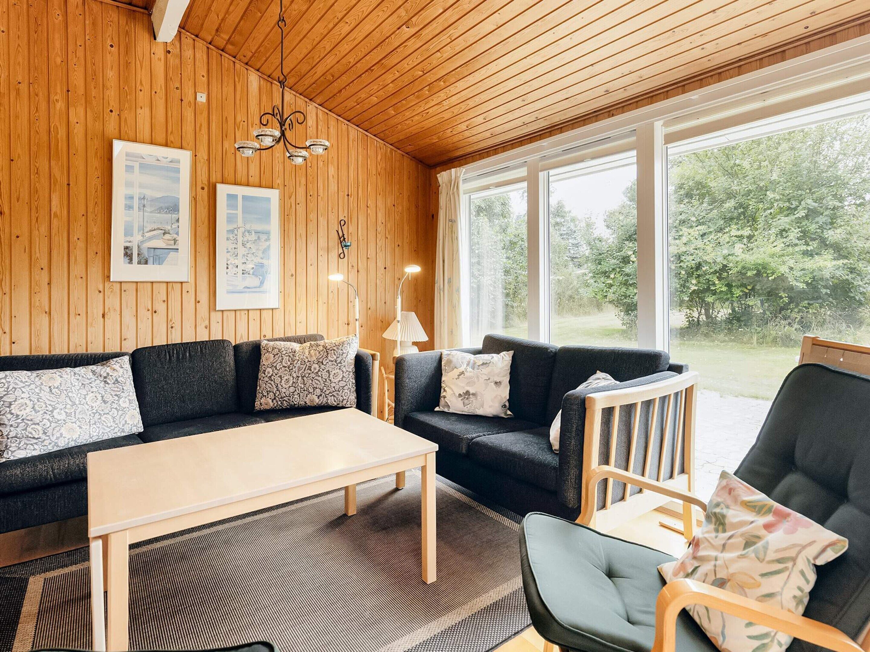 A cozy room with wooden walls, a sofa set, a table, and a chair. There are two windows providing natural light and a view of greenery outside.