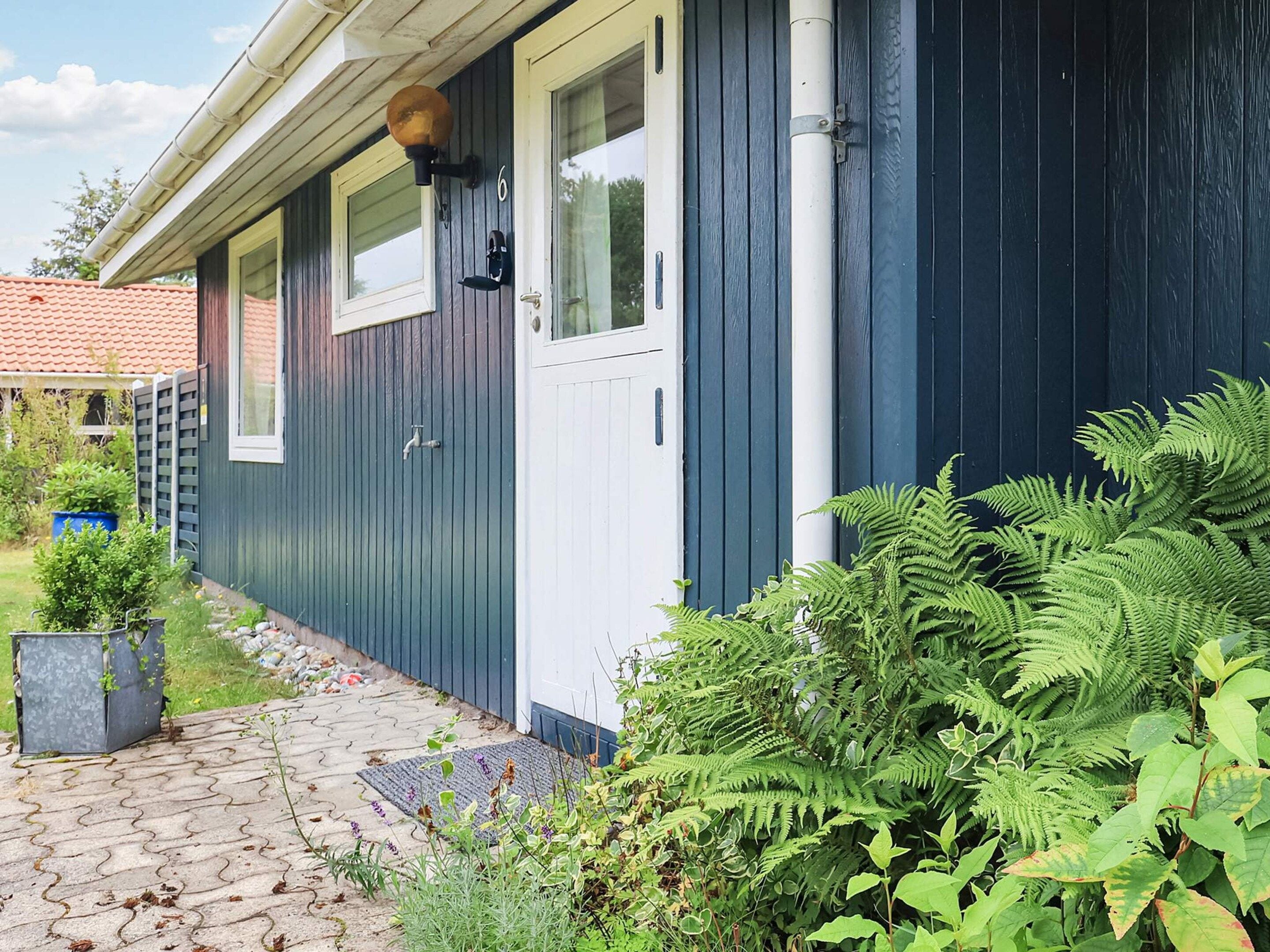A house with a dark blue exterior, a white door, and a window with a shutter. There is a garden with various plants and a paved walkway leading to the door.