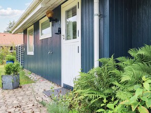A house with a dark blue exterior, a white door, and a window with a shutter. There is a garden with various plants and a paved walkway leading to the door. - 8 Person Holiday Home in Dannemare (Dannemare)