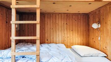 A bunk bed with a wooden ladder, a single bed with a white pillow, and a wall-mounted lamp.