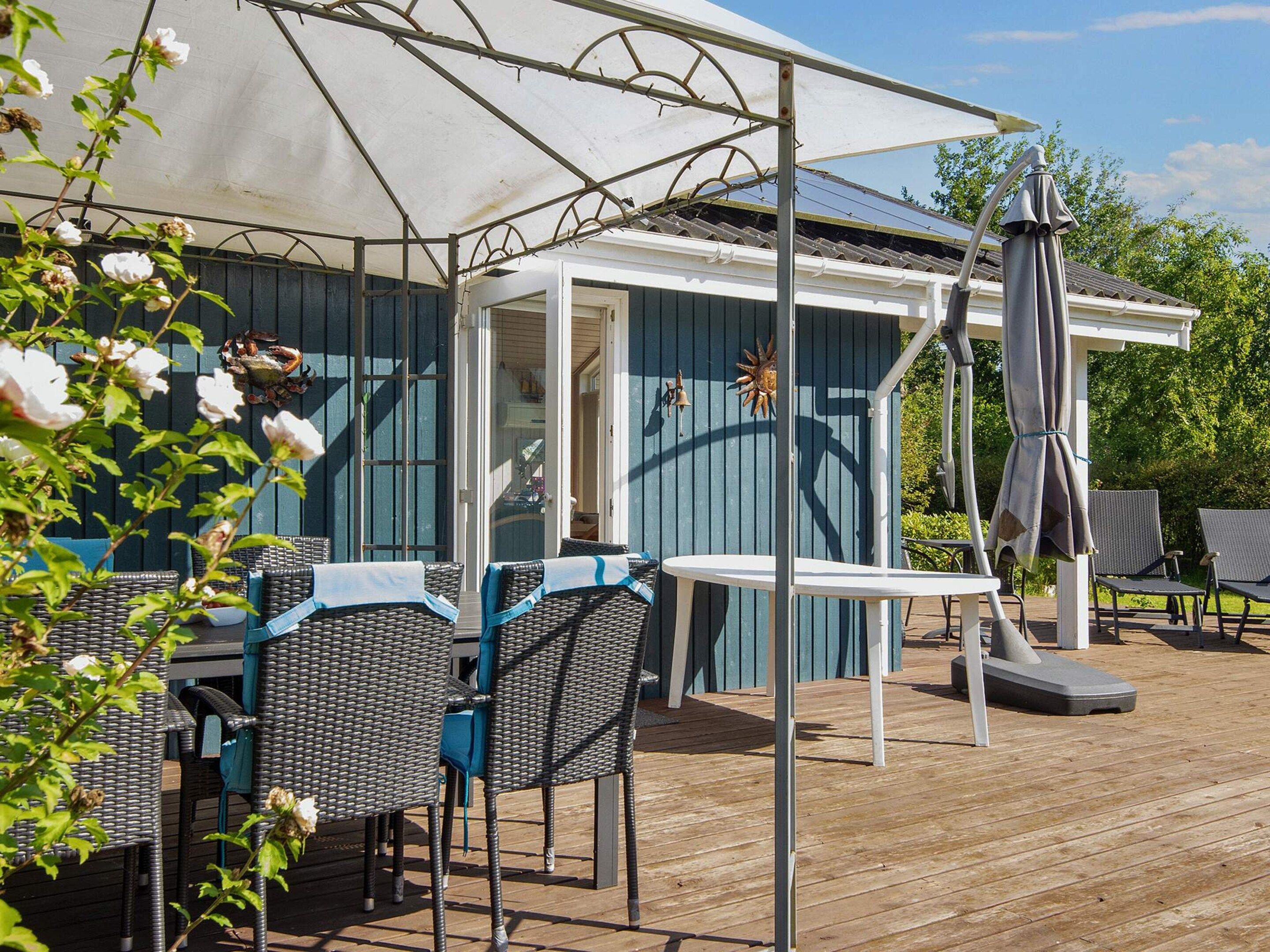 Een overdekt terras met rieten meubels en een tafel, een pergola met zonnescherm en uitzicht op een huis met een blauwe gevel.