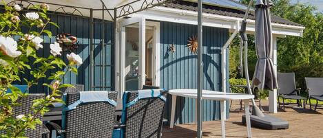 Een overdekt terras met rieten meubels en een tafel, een pergola met zonnescherm en uitzicht op een huis met een blauwe gevel.