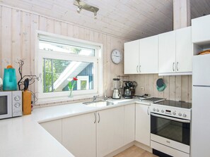 A compact kitchen with white cabinets, a stove, a sink, and a window with a view of a house. - Serene Holiday Home in Juelsminde near Beach (Juelsminde)