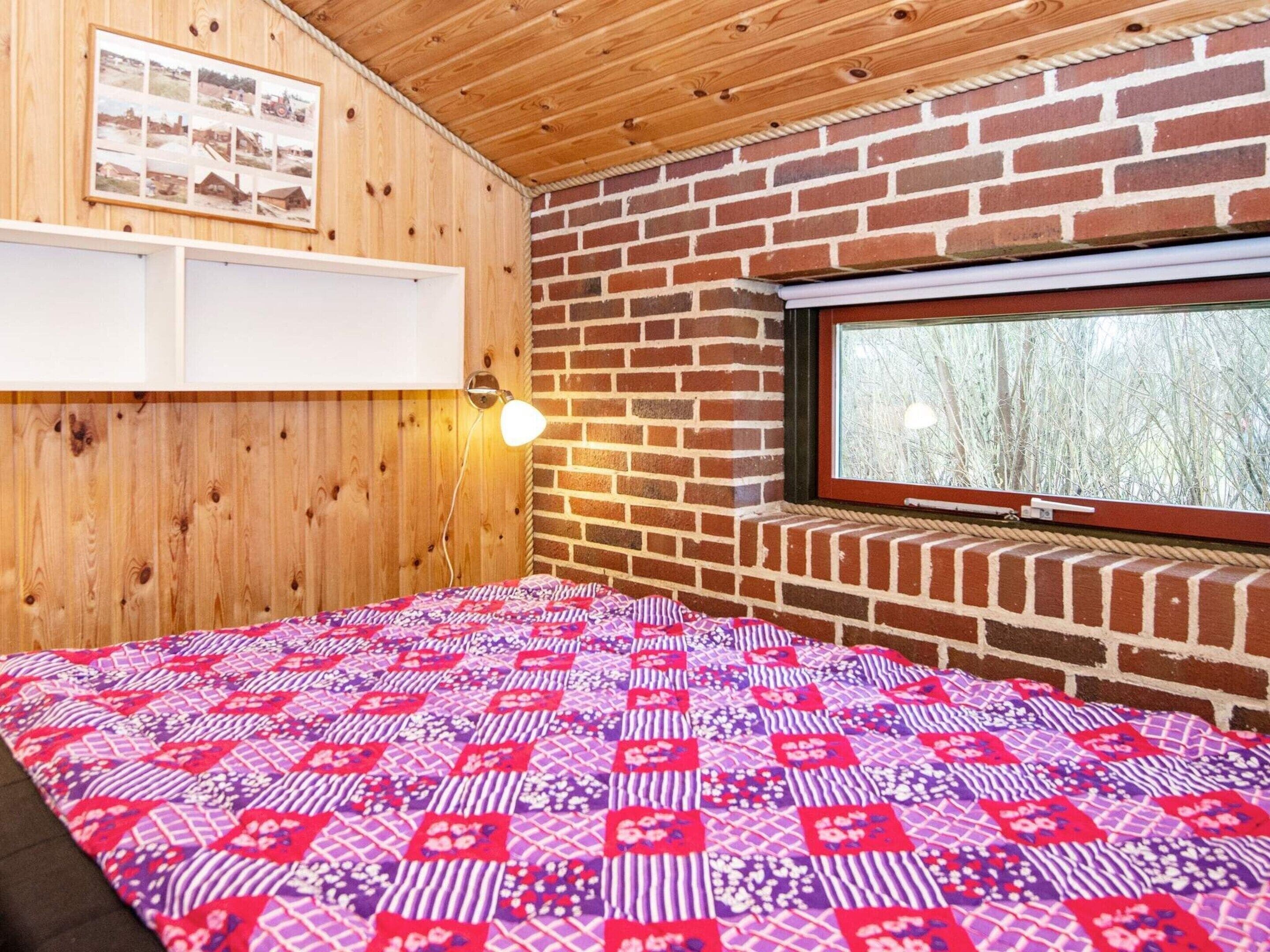 A room with a wooden ceiling, a brick wall, a window with a view of trees, a bed with a patterned bedspread, and a wall-mounted light fixture.