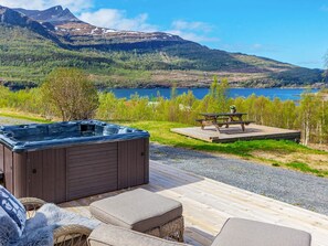 A hot tub, outdoor seating, and a picnic table on a deck with a lake and mountains in the background.
