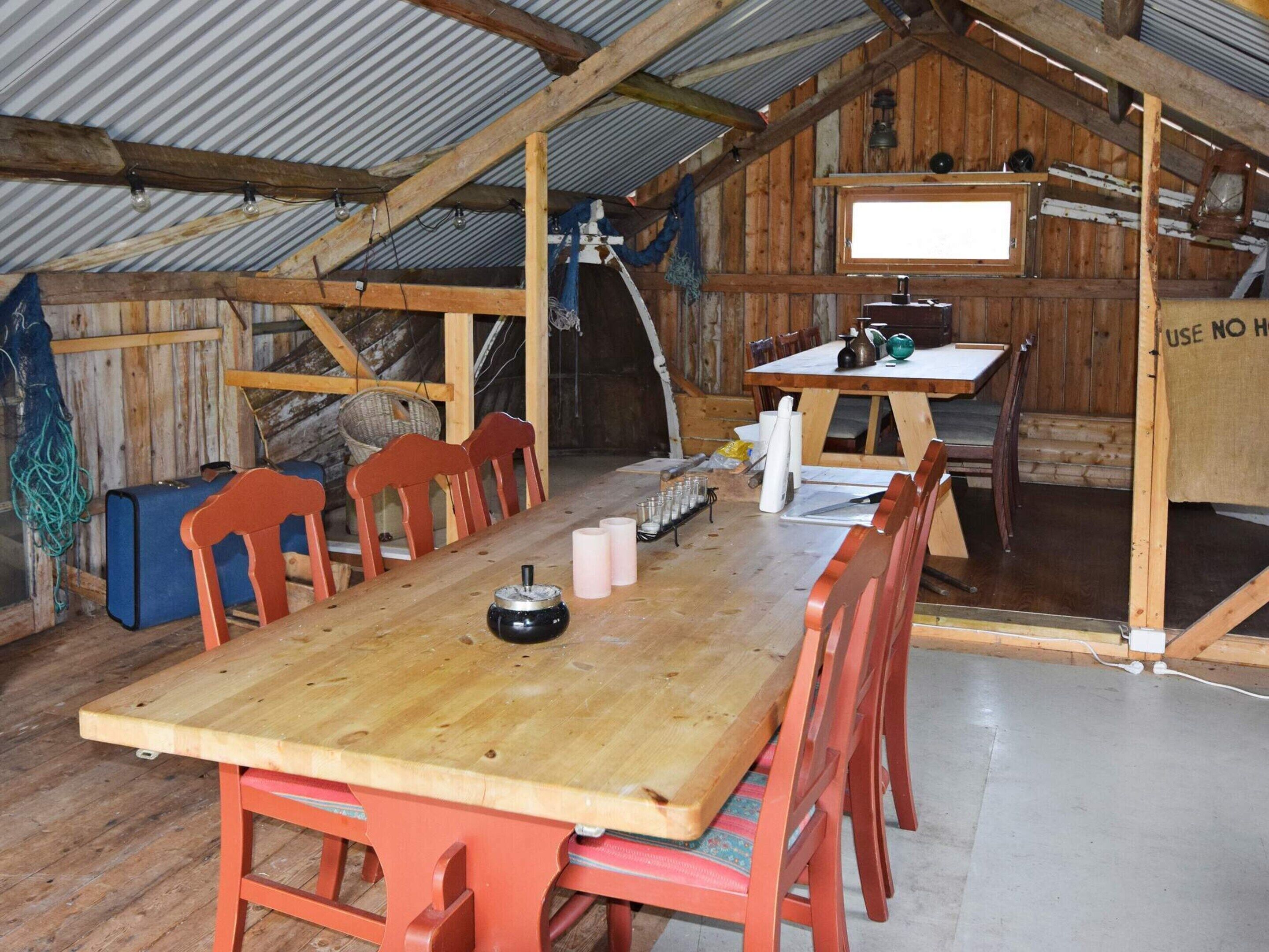 Un comedor rústico de madera con una mesa larga y sillas rojas, una pequeña cocina con una mesa y sillas, y un techo de láminas de metal corrugado.