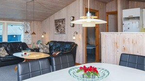 A cozy room with a wooden interior, a round dining table with a glass bowl of flowers, a black leather sofa with patterned cushions, and a kitchen area with a refrigerator.
