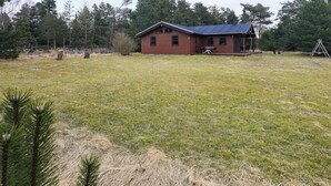 A single-story wooden cabin with a metal roof, surrounded by a grassy field and trees.