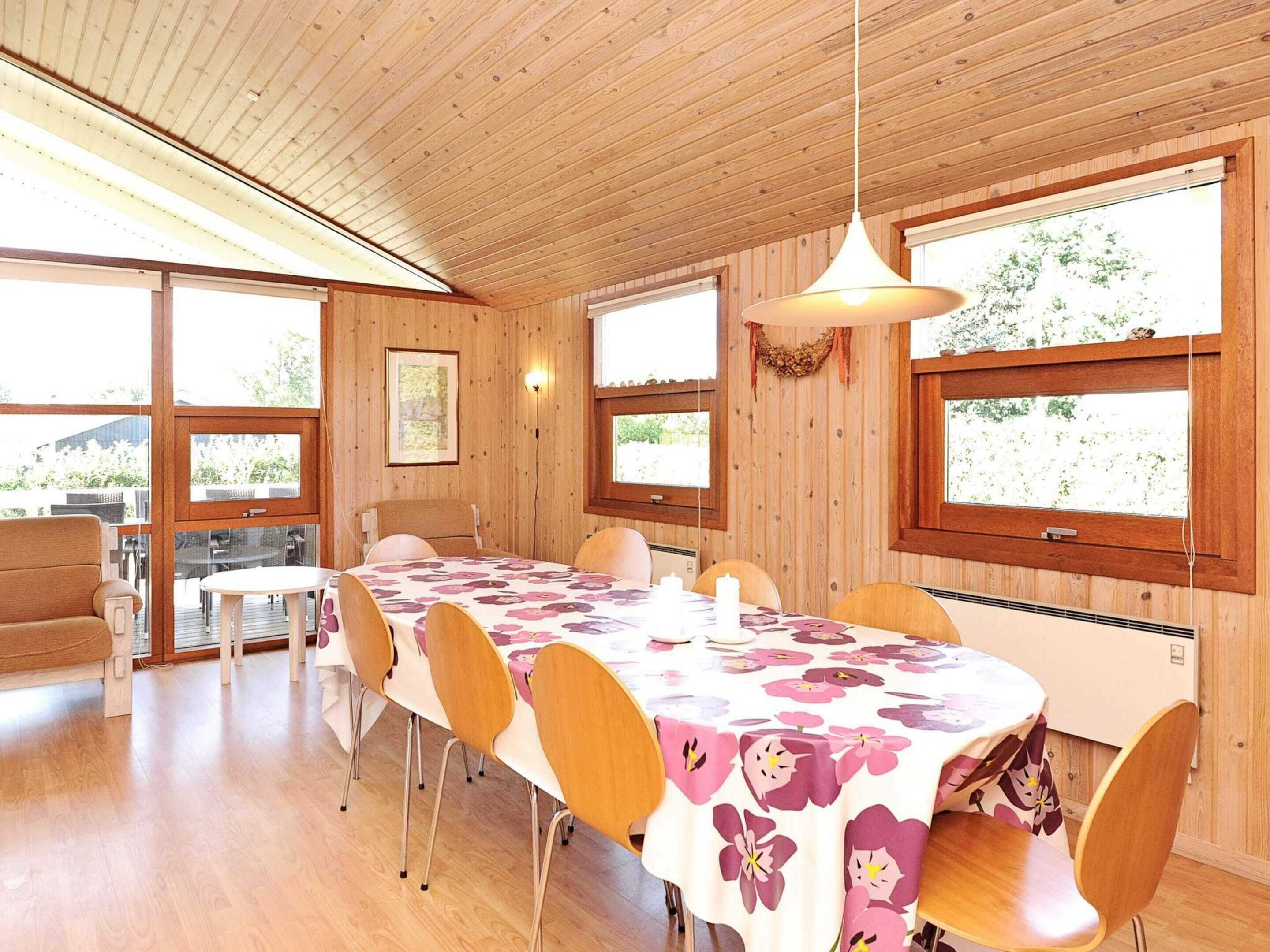 En matplats med ett långt bord täckt av en blommig duk, omgiven av trästolar, stora fönster och ett träpanelat tak.
