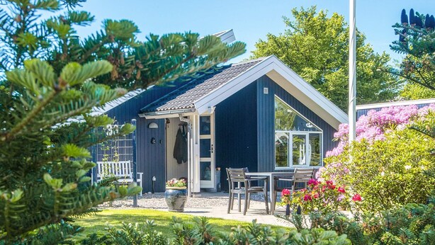 A house with a dark exterior, a white porch, and a garden with various plants and flowers. - Captivating Holiday Home in Ulfborg near Sea (Ulfborg)