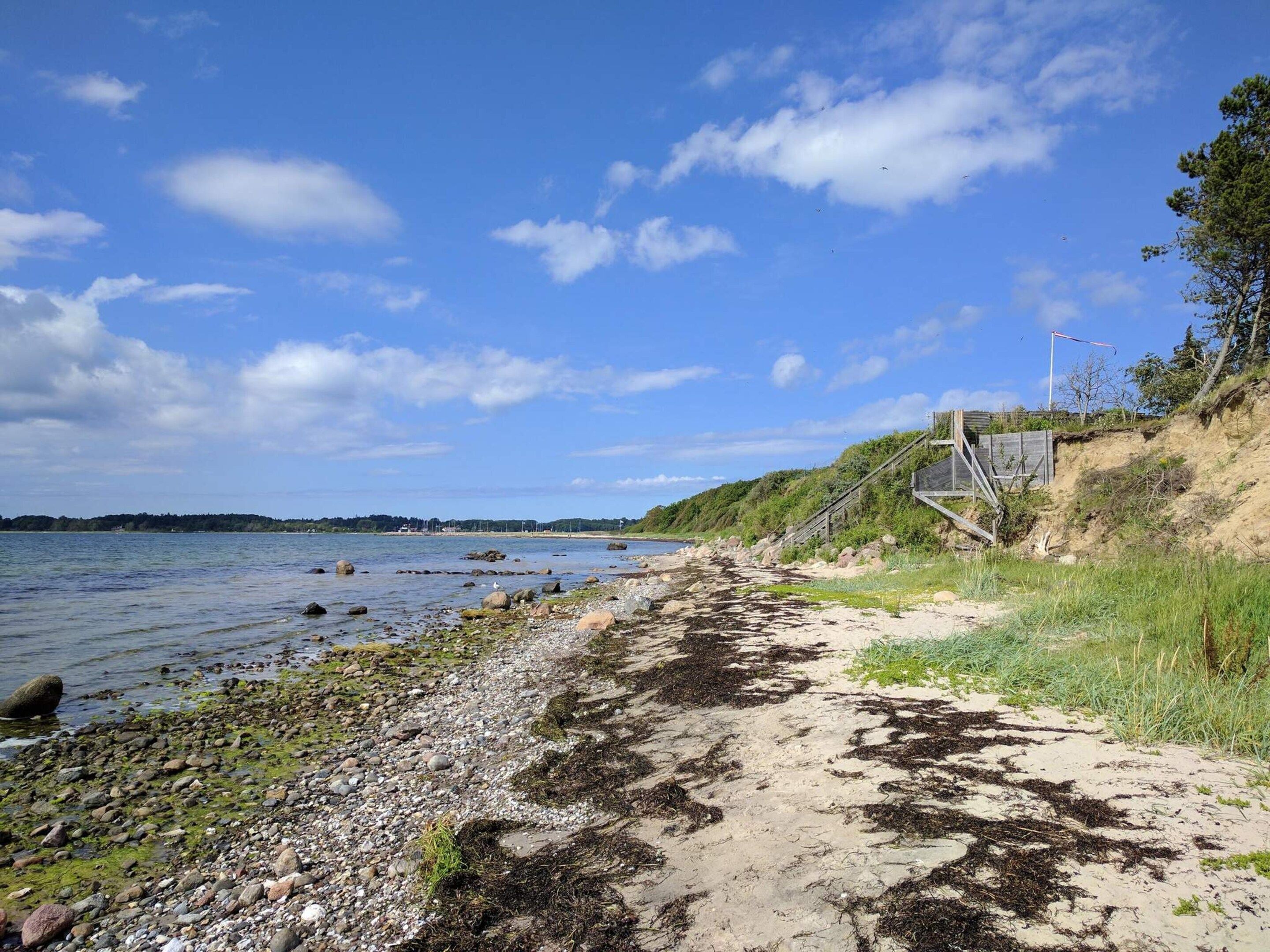 A coastal path with a sandy and rocky shoreline, a wooden staircase leading down to the beach, and a clear blue sky above.