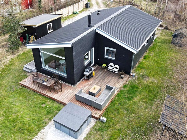 A modern house with a deck, outdoor seating, and a large window. - Woodland Holiday Home in Jægerspris with Hot Tub (Jægerspris)