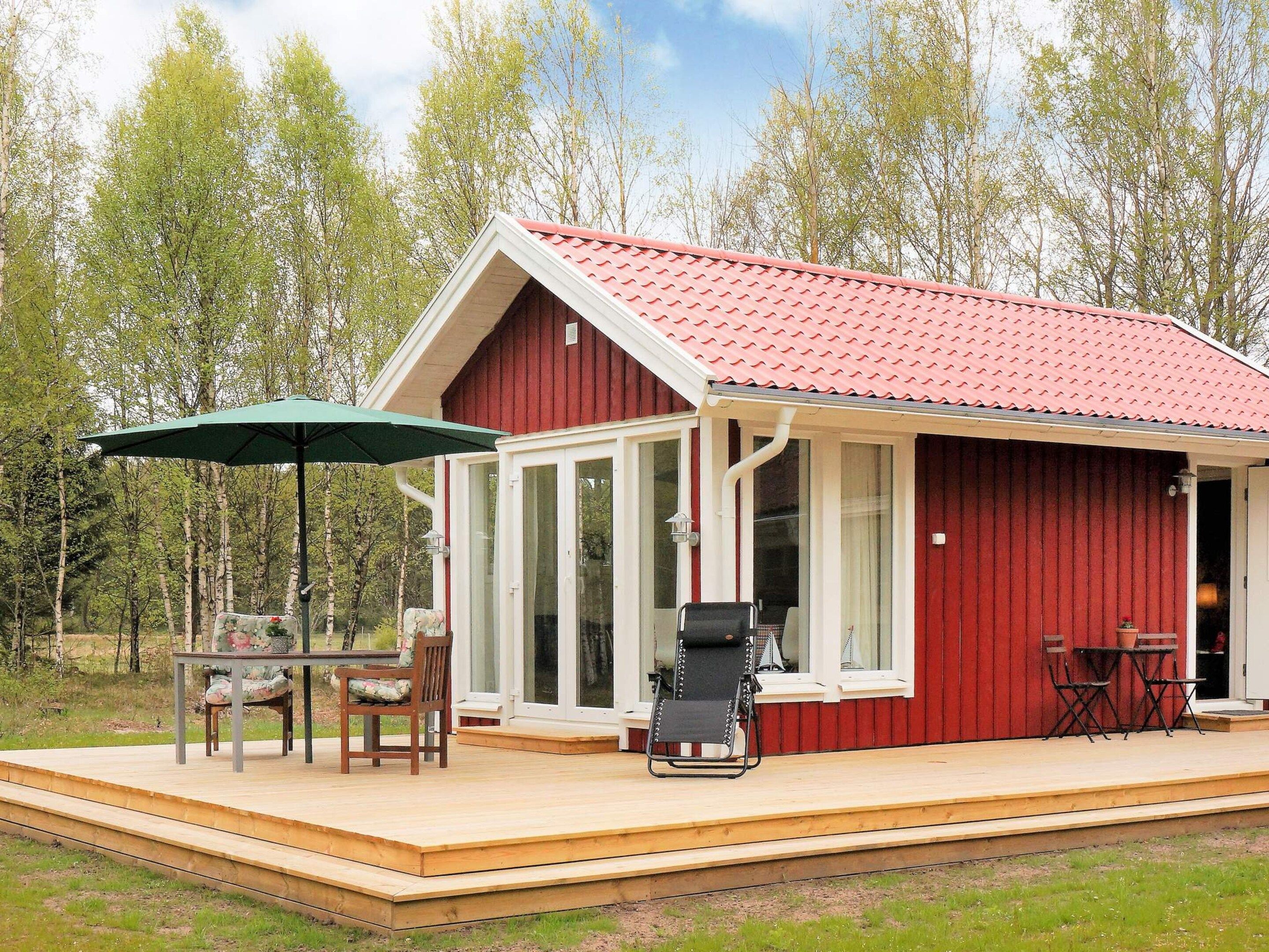 Een rode houten blokhut met een overdekt terras, een tafel met stoelen en een barbecue.