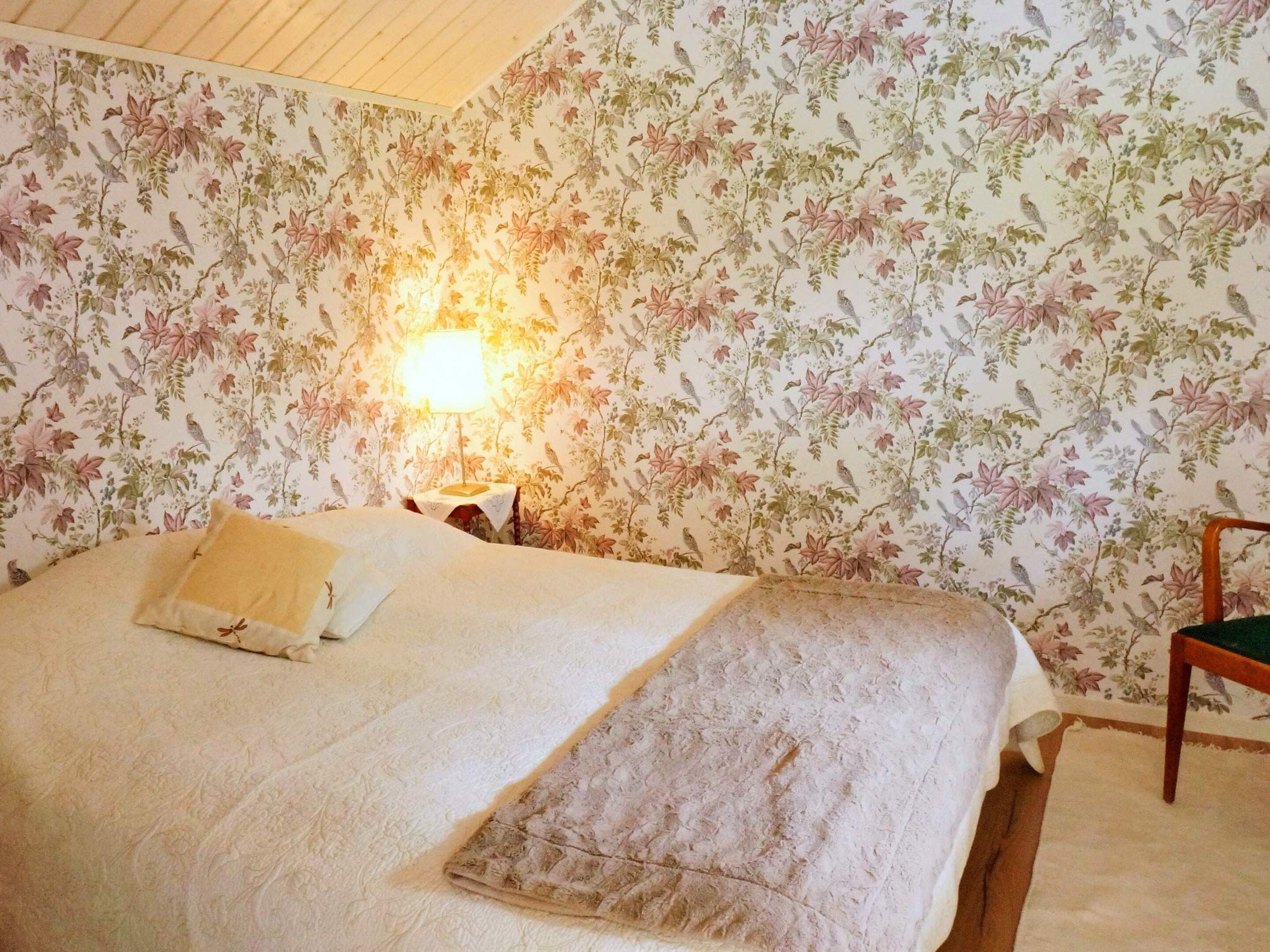 Un dormitorio con papel tapiz floral, una cama con colcha blanca, una mesita de noche con lámpara y una silla.