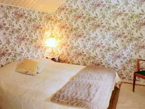 A bedroom with floral wallpaper, a bed with a white bedspread, a nightstand with a lamp, and a chair. - 6 Person Holiday Home in Heberg (Heberg)
