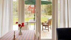 Ett rum med ett rutigt bord, en vas med röda blommor, ett ljus och en utsikt över en träveranda och ett rött hus genom en glasdörr.