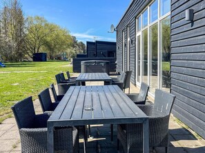 Eine Terrasse im Freien mit Holztisch und Korbstühlen, eine Glastür zu einem überdachten Bereich sowie ein Spielplatz im Hintergrund.
