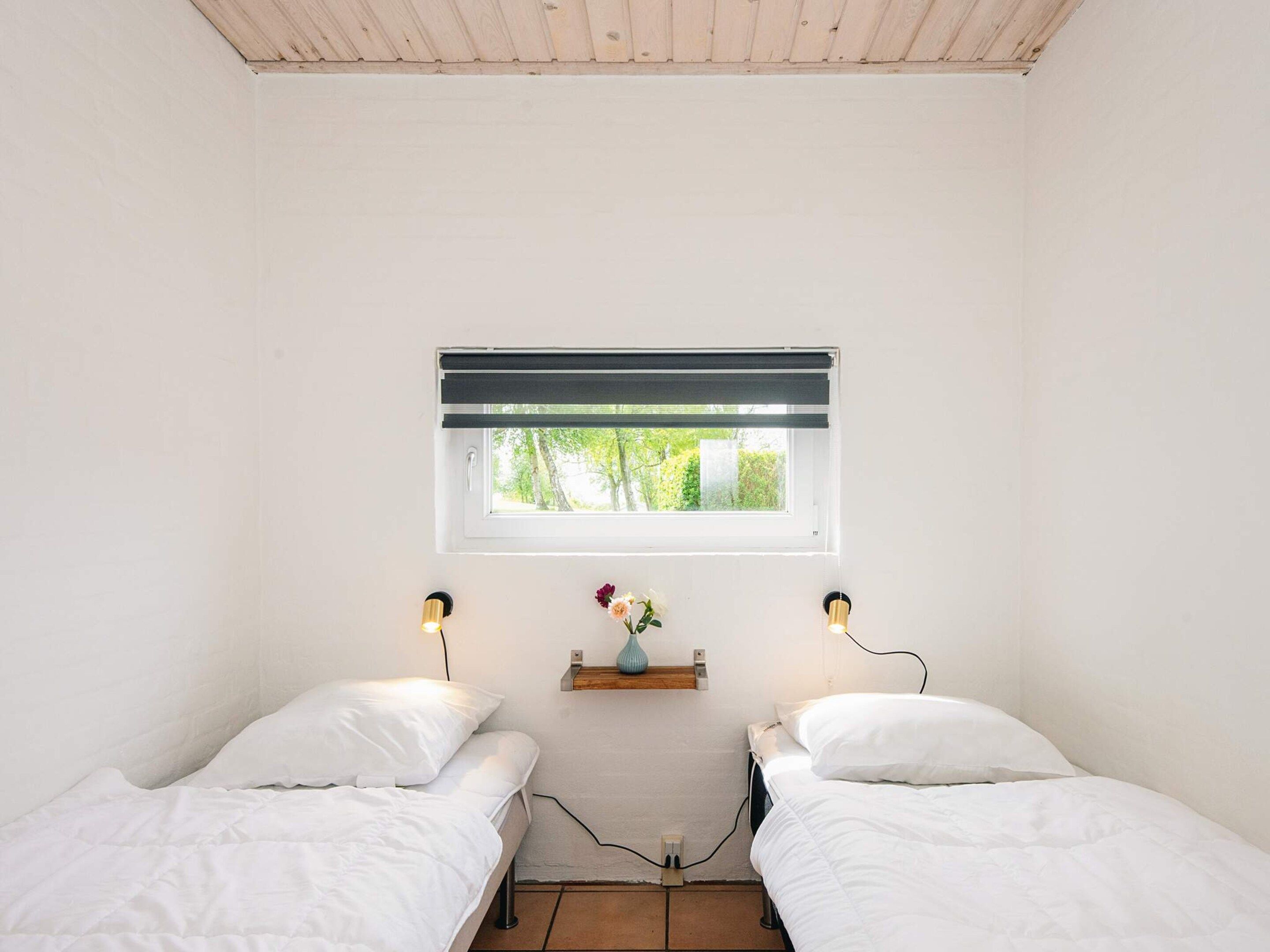 A bedroom with two beds, a window with blinds, a shelf with a vase, and wall-mounted lamps.