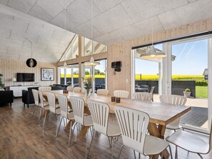 A modern dining area with a long wooden table and white chairs, a television on the wall, and large windows offering a view of the outdoors.