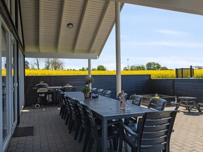 An outdoor patio with a long table and chairs, a grill, and a view of a yellow flower field. - 5 Star Holiday Home in Bogense (Bogense)