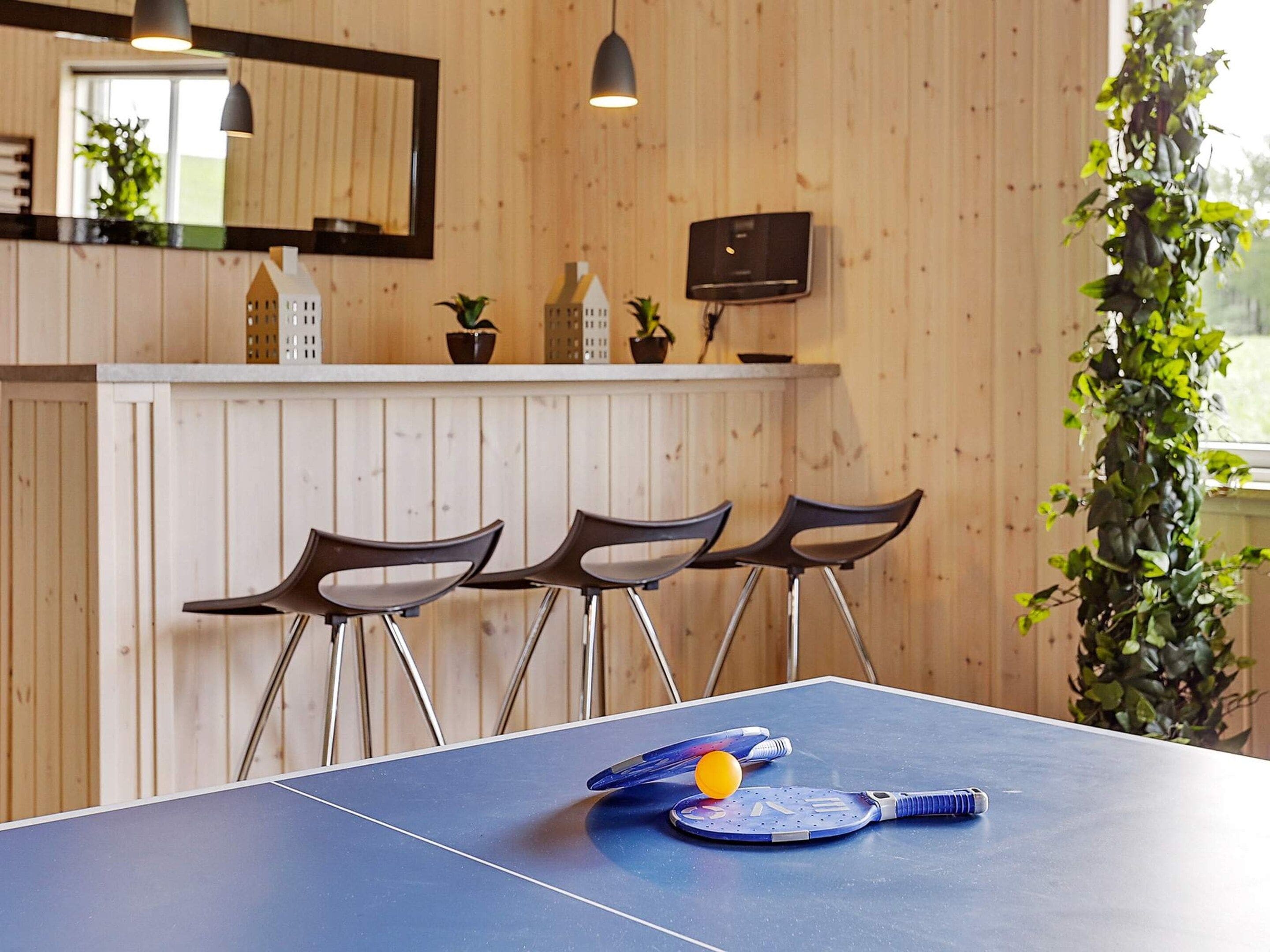A ping pong table with two paddles and a ball, three chairs, a wooden wall with a mirror, and a potted plant.