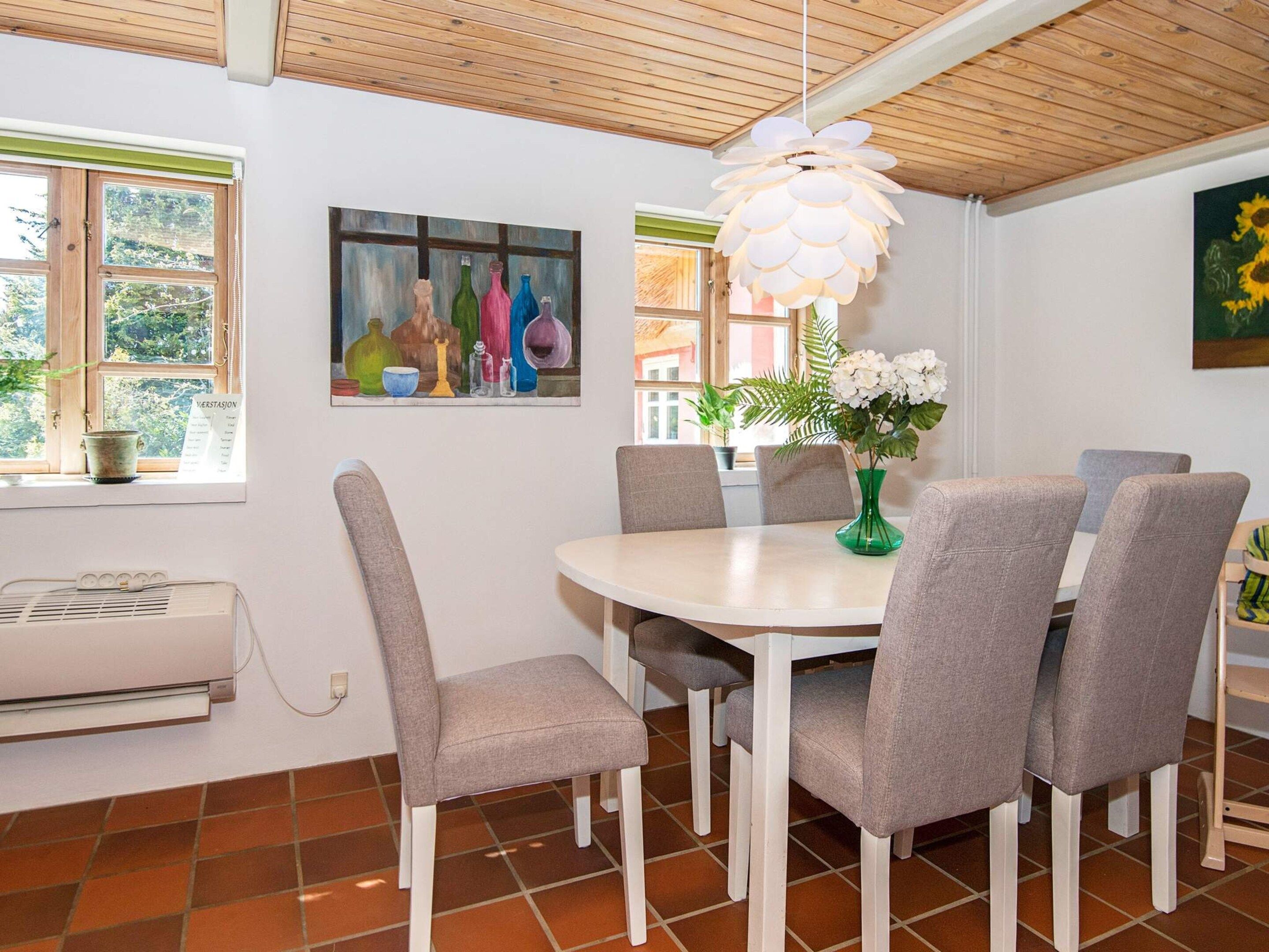 A dining area with a round table, grey upholstered chairs, a green vase with white flowers, a painting on the wall, and a window with a view of greenery.