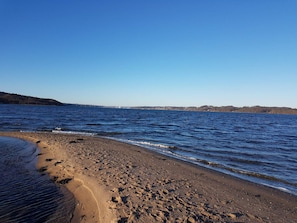 Uma praia com águas azul-claras, areal arenoso e uma ponte ao longe.