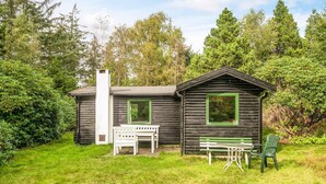 Una cabaña de troncos con chimenea, un banco y una mesa con sillas en una zona verde rodeada de árboles.