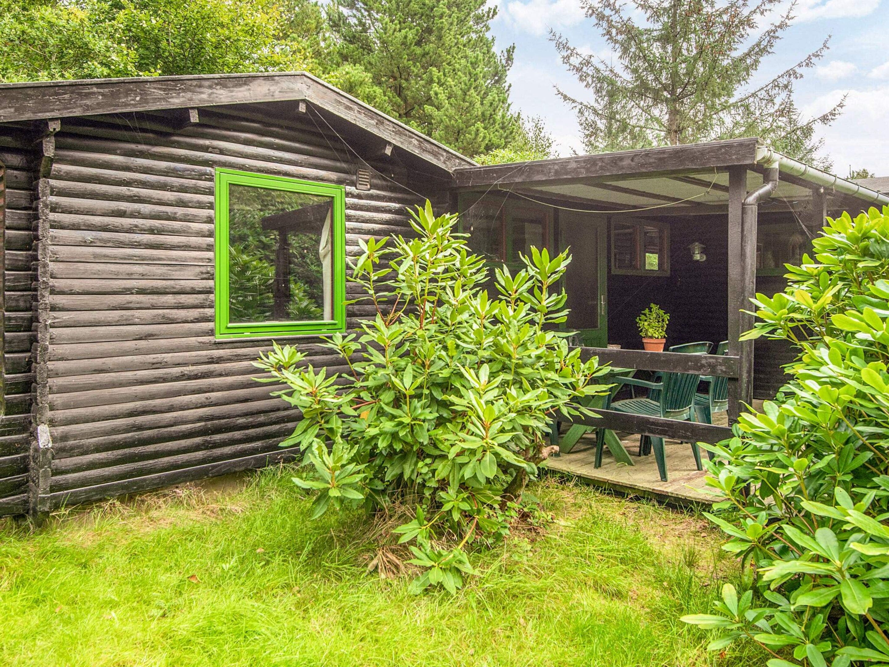 A rustic cabin with a green-framed window, a covered porch with chairs, and a small table.