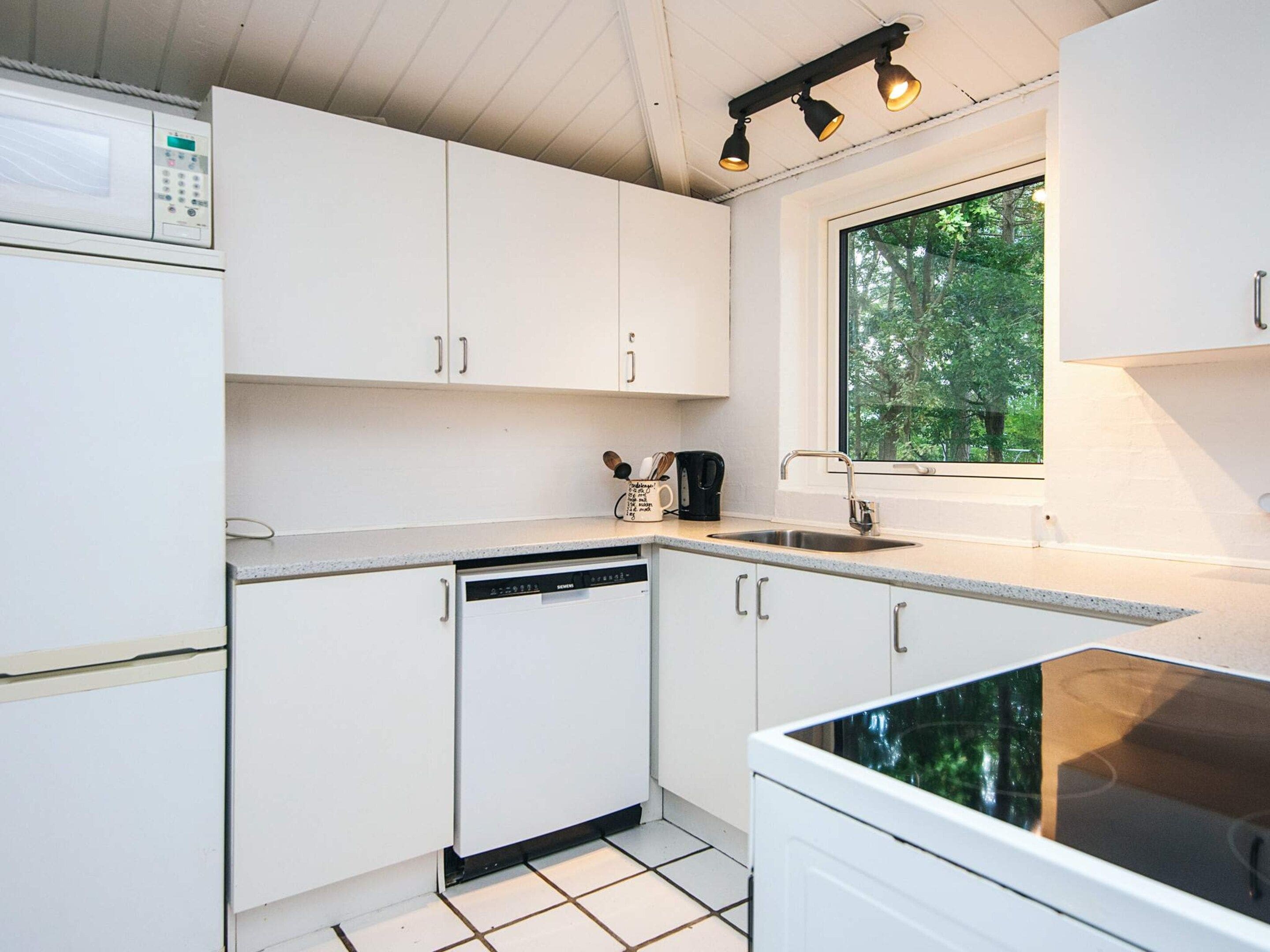 Cocina compacta con gabinetes blancos, lavavajillas, microondas y ventana con vistas a zonas verdes.